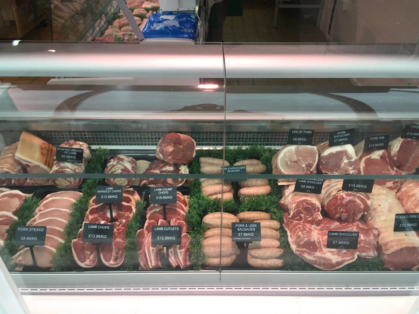 The chilled counter at Olney Butchers: joints of pork and lamb, chops, cutlets and two trays of sausages, each with a black ticket, set on green garnish